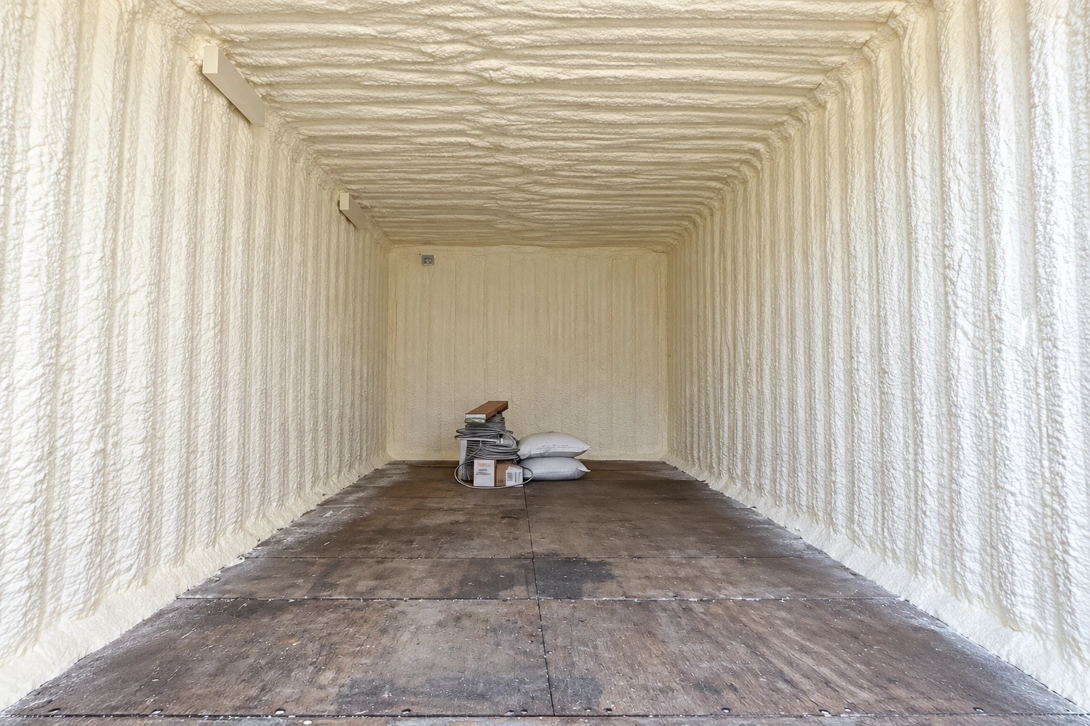 A narrow room with cream-colored spray foam covering its walls and ceiling, leaving the concrete floor exposed.