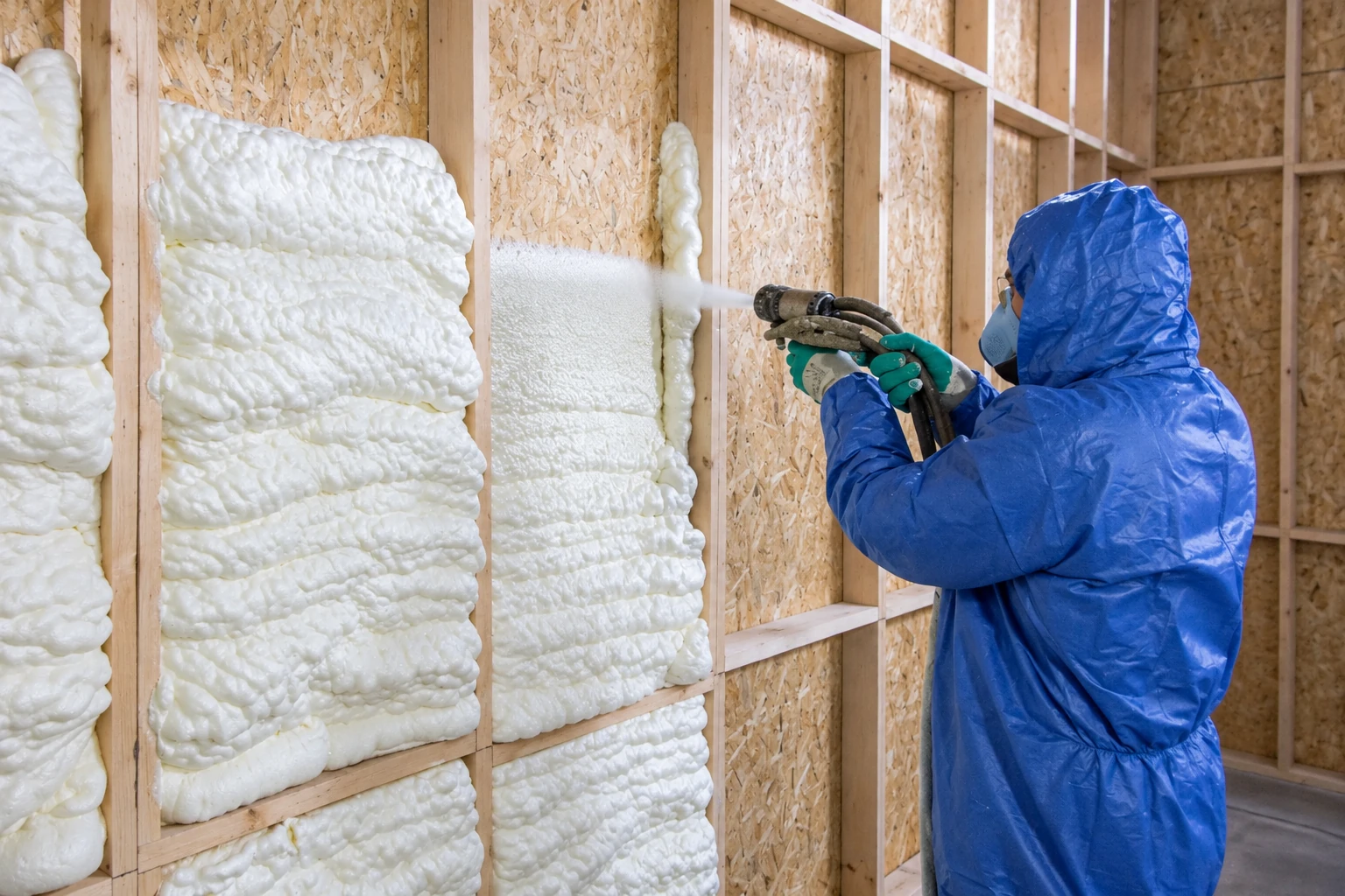 A worker in blue protective gear sprays white foam insulation into exposed wall framing.