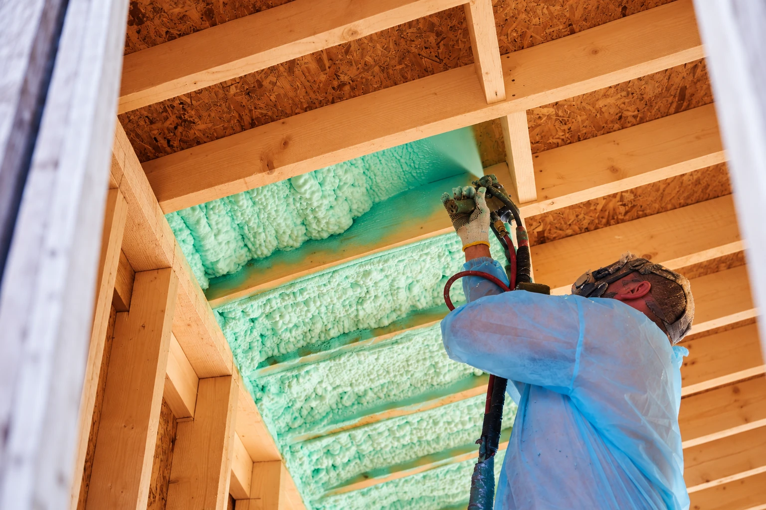 A worker in protective clothing sprays green foam insulation between exposed wooden roof rafters.