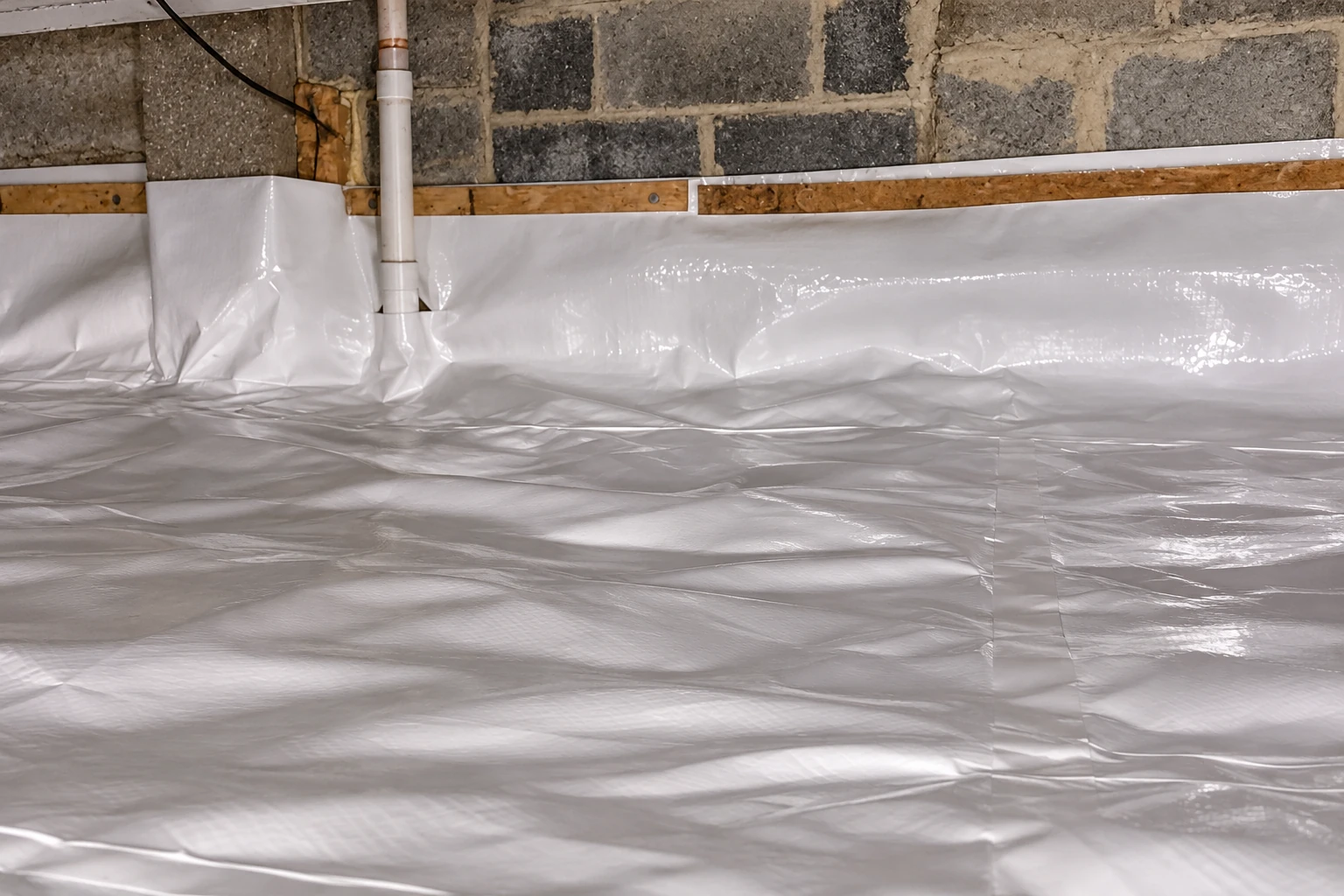 A white plastic vapor barrier covers the floor and lower walls of a basement with exposed block walls and a pipe.