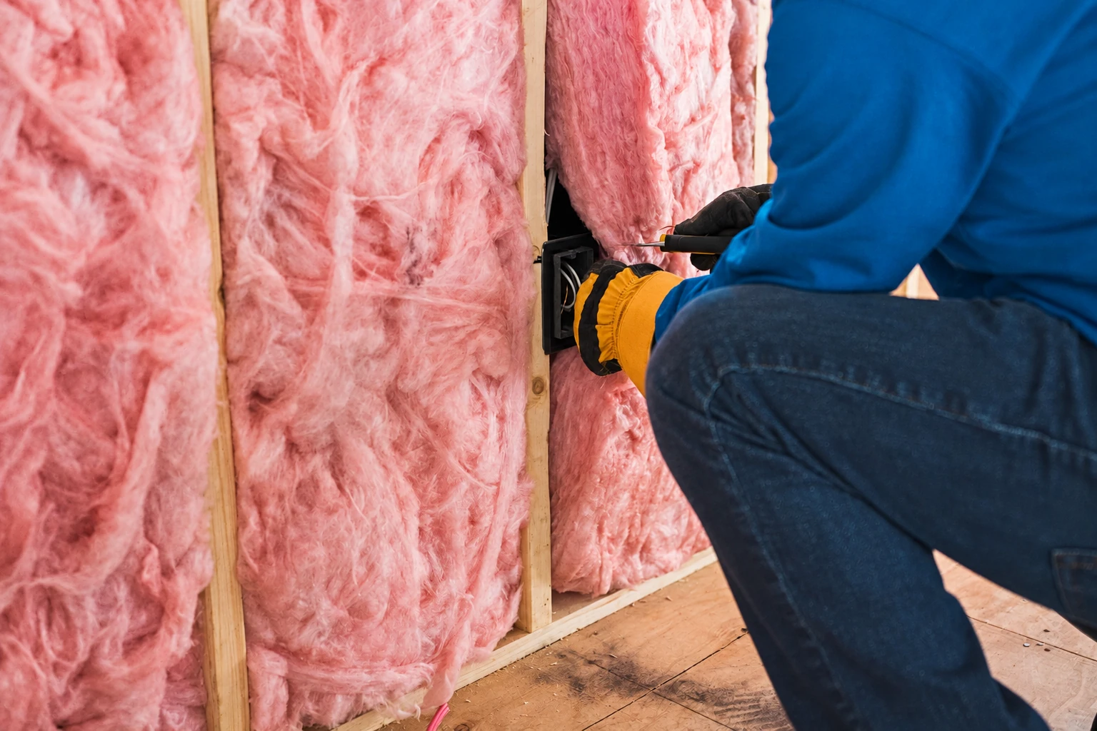 A worker in gloves fits pink fiberglass batts between wooden wall studs beside an electrical box.