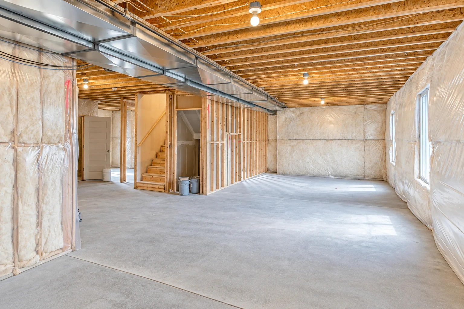 Unfinished basement with white insulation covered in clear plastic along the walls beneath exposed ceiling joists.